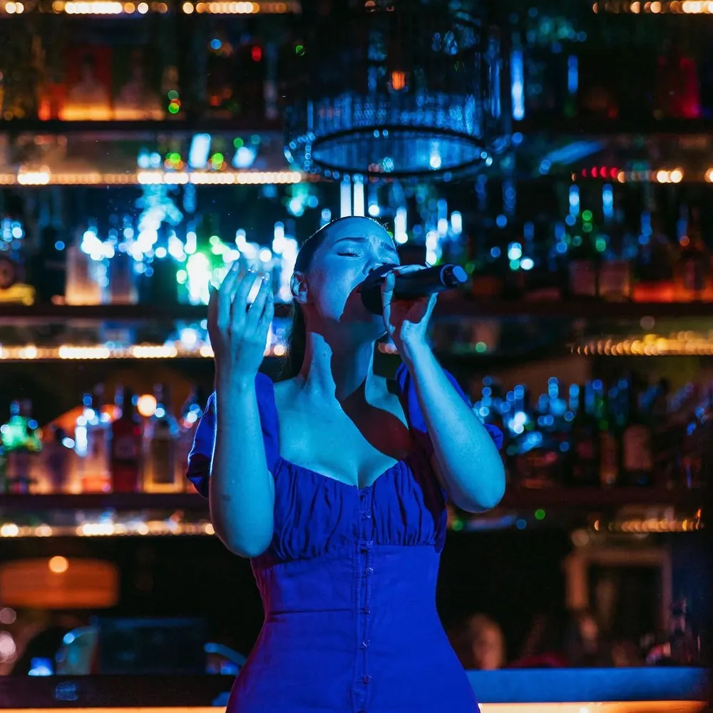 Rocío cantando con vestido azul delante de una barra con botellas iluminadas