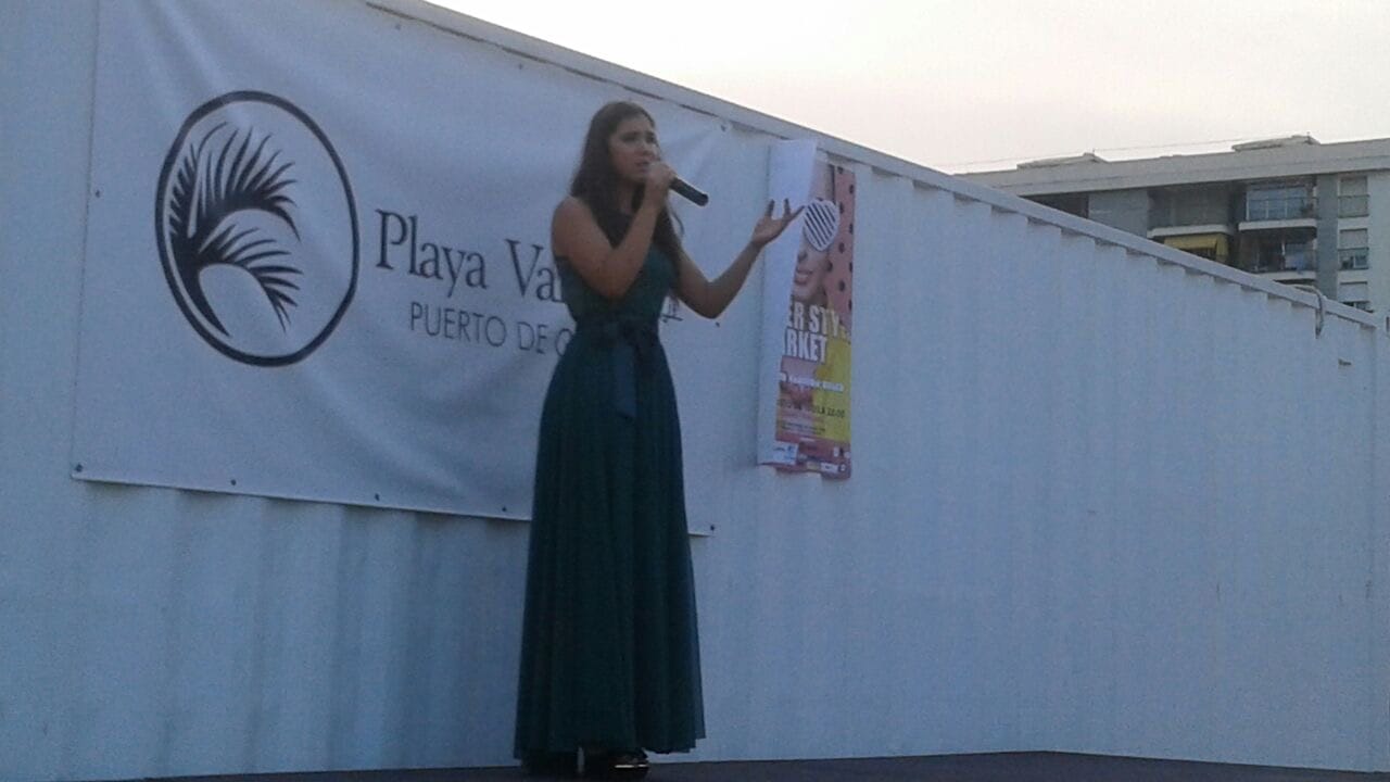 Rocío cantando al aire libre con micrófono y vestido largo verde en un escenario en el puerto, junto al mar.