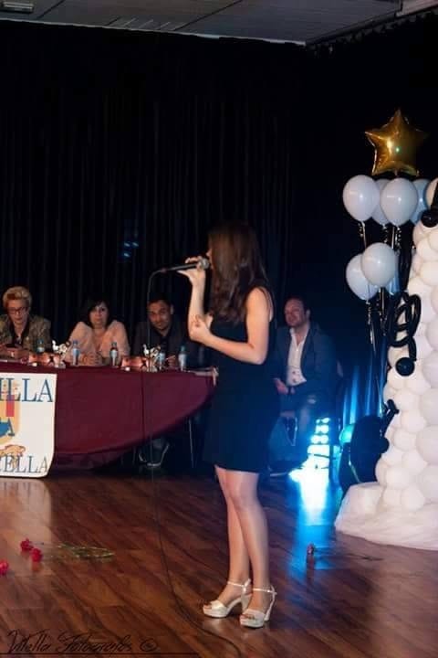 Rocío cantando con vestido negro y micrófono sobre un escenario de madera, ante una mesa de jurado.