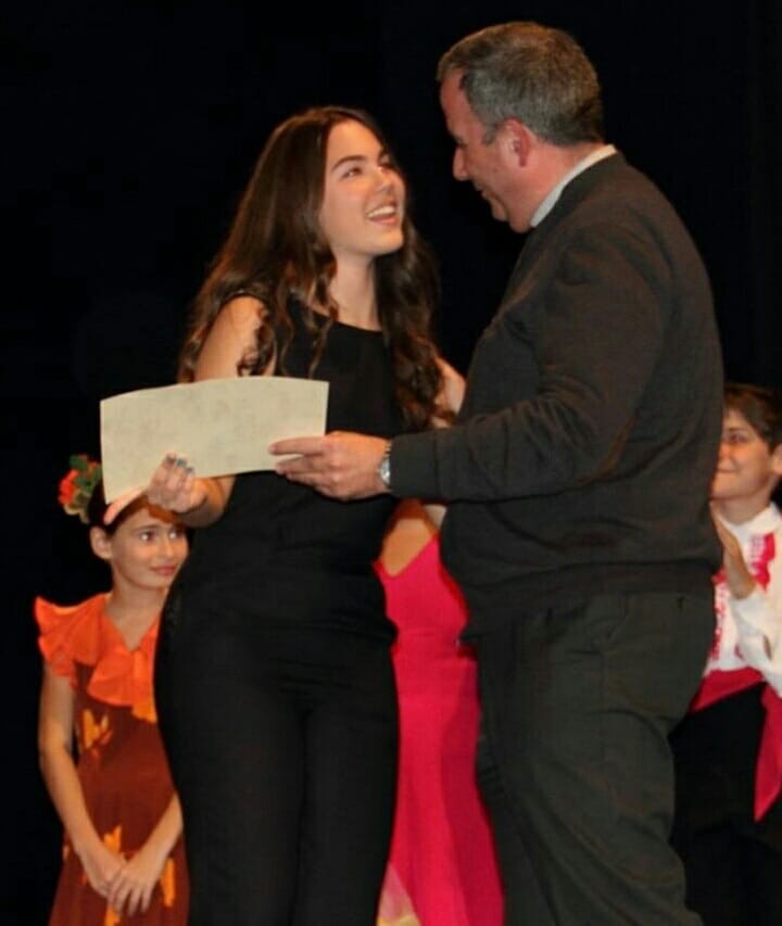 Rocío, vestida de negro, recibiendo sonriente un diploma de manos de un organizador en la entrega de premios de un certamen.