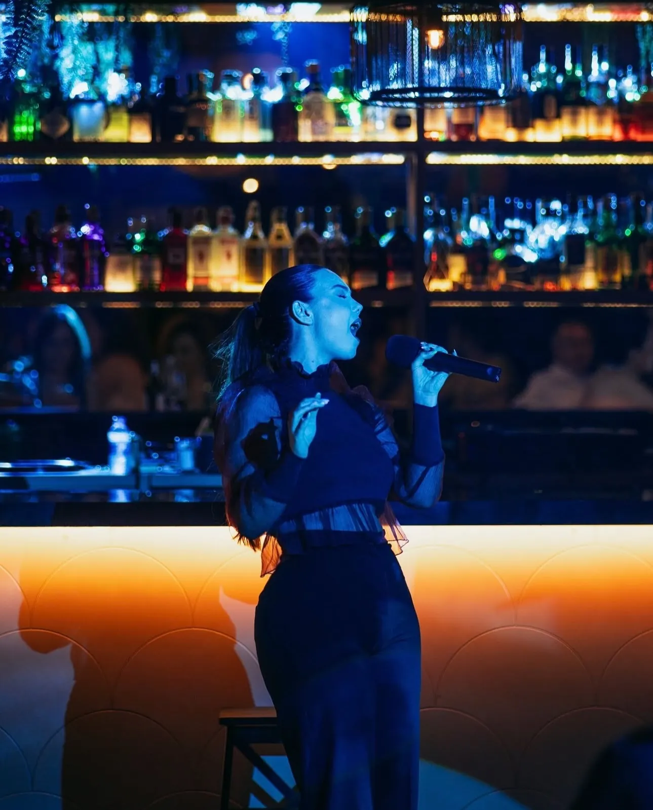 Rocío cantando de pie delante de una barra con botellas iluminadas, con top negro transparente